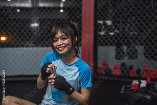 A tired but smiling twenty something athlete sits on the ring cage floor and beams for the camera as she feels energized by the sports drink she is holding in her hands. photo