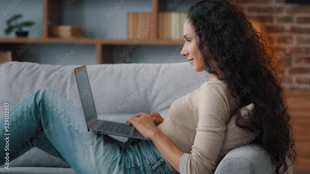 Side view smiling writer freelancer woman sitting on couch at home typing laptop keyboard writing article chatting with friends in online computer app web social network working remote distant job Side view smiling writer freelancer woman sitting on couch at home typing laptop keyboard writing article chatting with friends in online computer app web social network working remote distant job