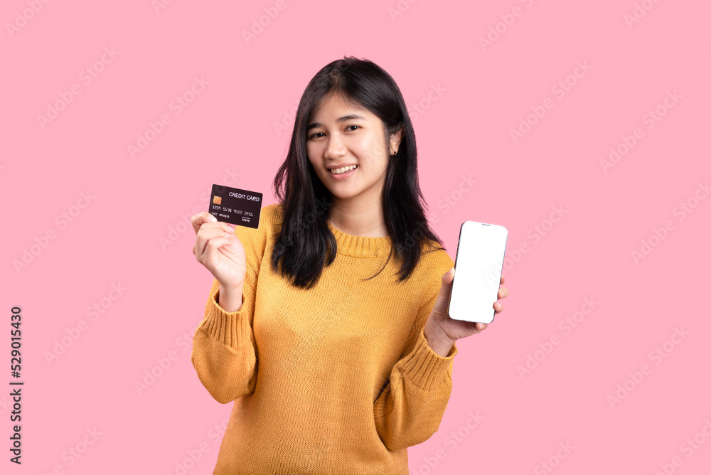 Portrait of a delighted woman standing on a yellow background. holding a cell phone showing a plastic credit card - obrazy, fototapety, plakaty 