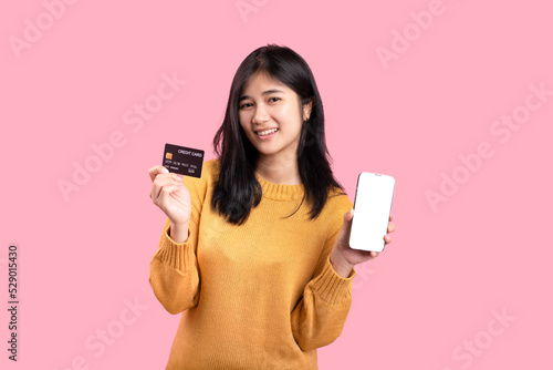 Portrait of a delighted woman standing on a yellow background. holding a cell phone showing a plastic credit card