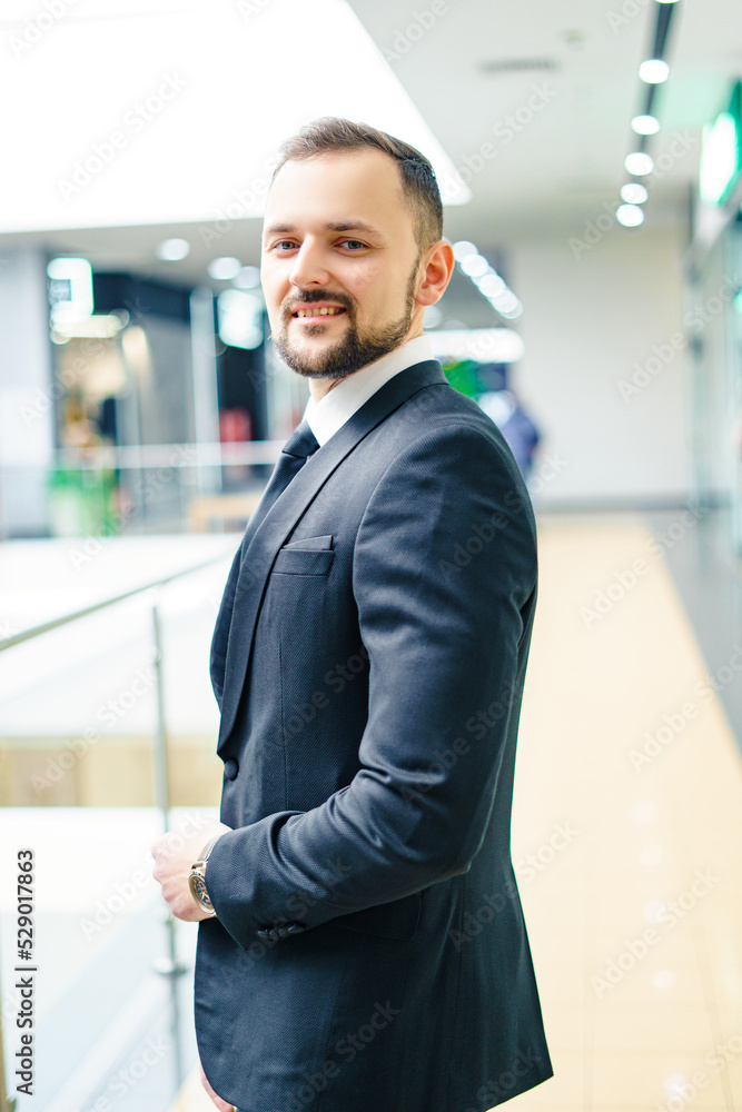 A young man with a beard dressed in a classic business suit. Young businessman in a mall wearing a black suit and black tie A young man with a beard dressed in a classic business suit. Young businessman in a mall wearing a black suit and black tie