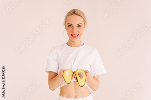 A smiling woman holds an avocado and looks at the camera, the girl is on proper nutrition and on a diet. White isolated background. Nutritionology. Place for text