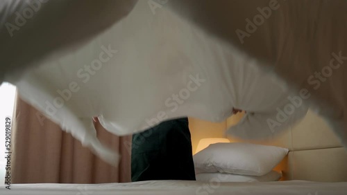 Chambermaid in uniform adjusts white blanket in hotel room looking in camera. Young woman makes bed and takes care of comfort for guests slow motion