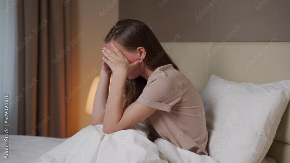 Young woman feels depressed sitting on bed in hotel room near bright lamp. Brunette lady puts hands on face trying to handle emotions in evening Young woman feels depressed sitting on bed in hotel room near bright lamp. Brunette lady puts hands on face trying to handle emotions in evening