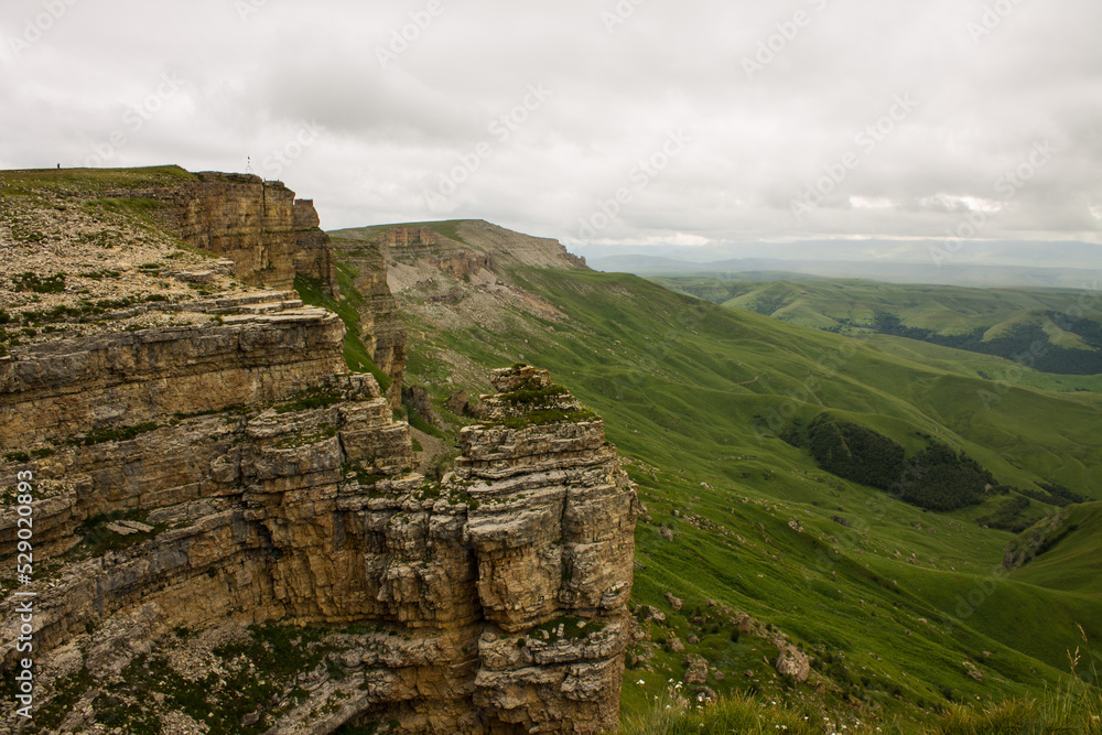 Naklejka premium Panoramic view of the canyon with green hills blurred by misty haze to the horizon and steep high cliffs of the Bermamyt plateau in Karachay-Cherkessia