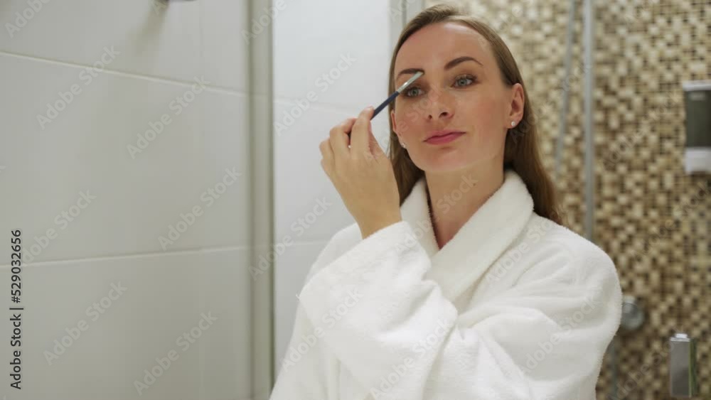 Young woman paints brows looking in mirror in hotel bathroom in morning. Female guest in white robe enjoys doing makeup after waking up closeup Young woman paints brows looking in mirror in hotel bathroom in morning. Female guest in white robe enjoys doing makeup after waking up closeup