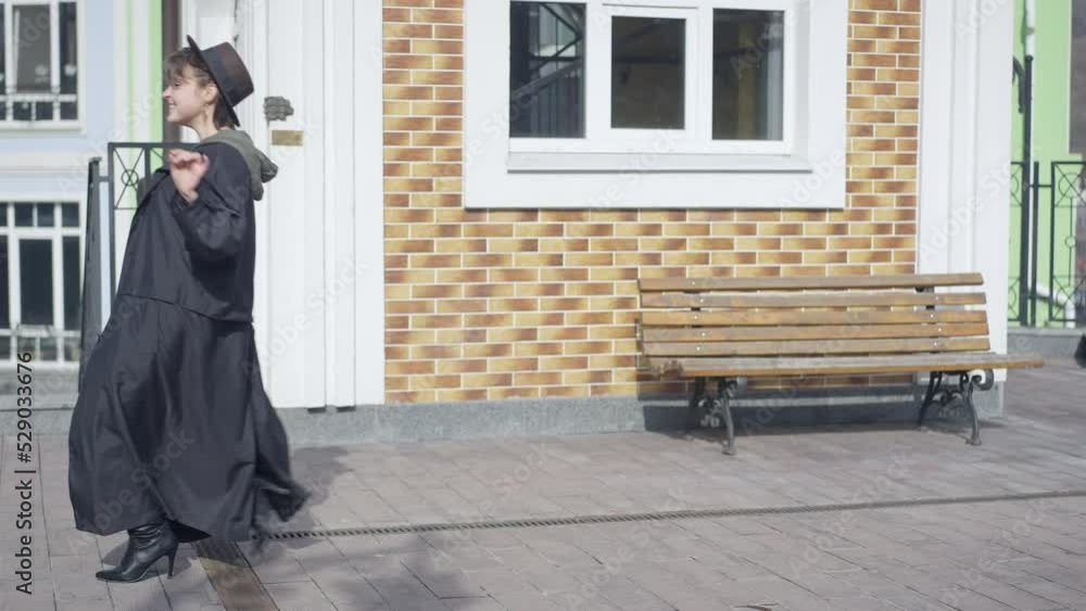 Funny carefree young woman in cloak and high-heels making steps looking at camera smiling. Wide shot side view of cheerful Caucasian slim lady in hat grimacing having fun on city street Funny carefree young woman in cloak and high-heels making steps looking at camera smiling. Wide shot side view of cheerful Caucasian slim lady in hat grimacing having fun on city street