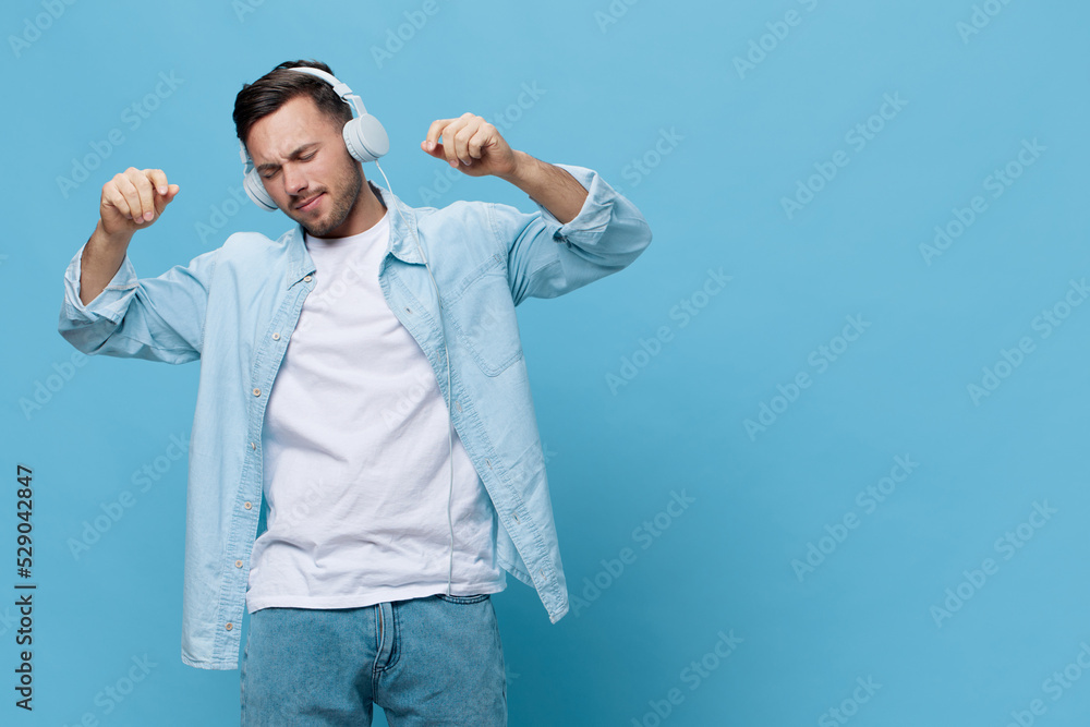 Fototapeta premium Joyful attractive tanned handsome man in casual basic t-shirt headphones listen good playlist dance closing eyes posing isolated on blue studio background. Copy space Banner Mockup. Music concept