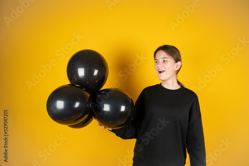 Pretty surprised brunette teengirl holding black balloons in her hand.Wearing black sweatshirt. Yellow studio background © Tatsiana