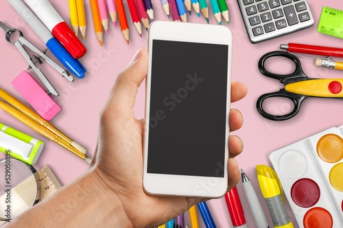 Hand holding a smartphone with a blank screen. Pink background with school supplies, accessories,