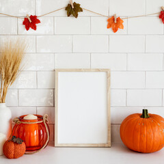 Picture frame mockup with orange pumpkins, vase of wheat, garland of leaves on wall tile background. Cozy home interior with autumn fall decor.