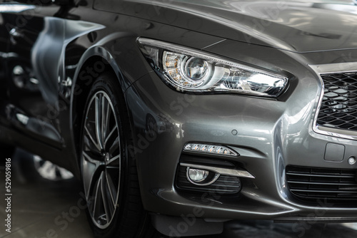 Front right modern headlight on luxury silver new car in automobile salon. Detail grey metallic bumper car with LED headlights and car wheel. Exterior automobile close up.