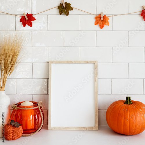Picture frame mockup with orange pumpkins, vase of wheat, garland of leaves on wall tile background. Cozy home interior with autumn fall decor.