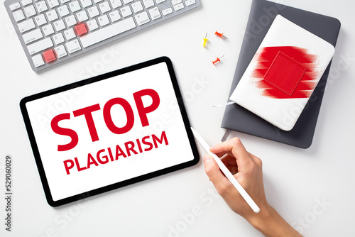 Stop plagiarism concept. Office desk table with keyboard, paper notebooks, tablet. Message STOP PLAGIARISM on tablet screen. photo