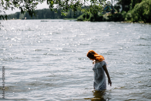 Fototapeta Naklejka Na Ścianę i Meble -  Woman in summer white dress stands on the seashore and looks at the horizon. Young beautiful girl standing in the water