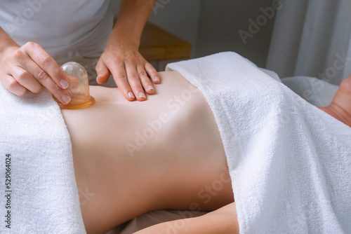 Spa procedure in a massage room, a woman patient lies relaxed on the table covered with a white terry towel, gentle hands of a female massage therapist massage her belly