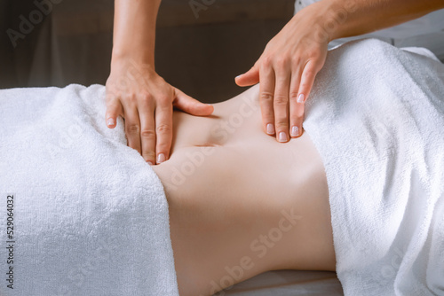 Spa procedure in a massage room  a woman patient lies relaxed on the table covered with a white terry towel  gentle hands of a female massage therapist massage her belly