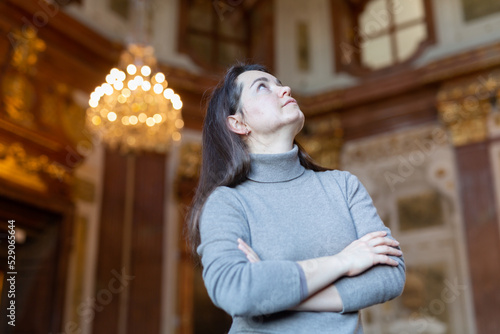 Photography Interested woman tourist admiring sumptuous interiors of medieval palace decorated with gold and antique chandelier in blurred background