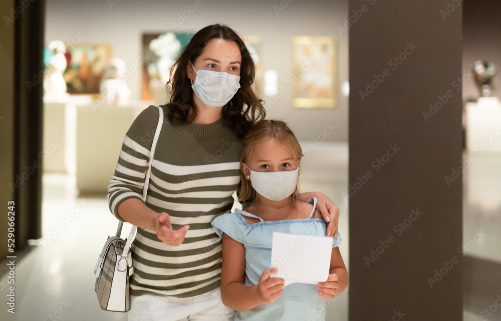 Woman with daughter in medical masks in museum of arts, looking at art objects