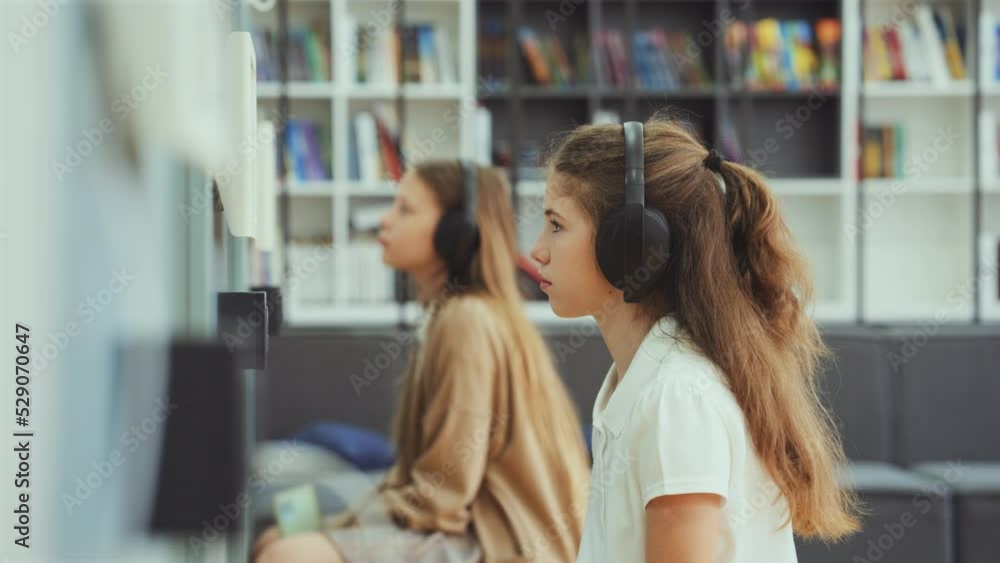Girls sitting in class wearing headphones and looking at device on wall, focus on child in front, bookshelves on background. Arc shot pupils studying at school. Concept of education Girls sitting in class wearing headphones and looking at device on wall, focus on child in front, bookshelves on background. Arc shot pupils studying at school. Concept of education
