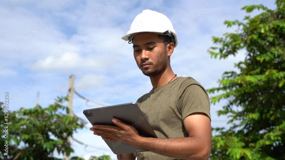 Dark brown skin engineer asian handsome man monitoring and working on tablet wearing white hard hat and green shirt with tree and high voltage power line in background. Dark brown skin engineer asian handsome man monitoring and working on tablet wearing white hard hat and green shirt with tree and high voltage power line in background.
