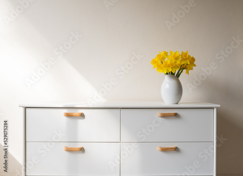 Yellow daffodils in white vase on side table against beige wall with light from window (selective focus)