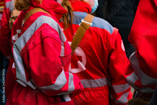 A crowd of emergency medic assistants in red uniforms stands on the street. Accident. Aid. Ambulance. Assistance. Assisting. Care. Civil. Crisis. Cure. Doctor. Emergency. Staff
