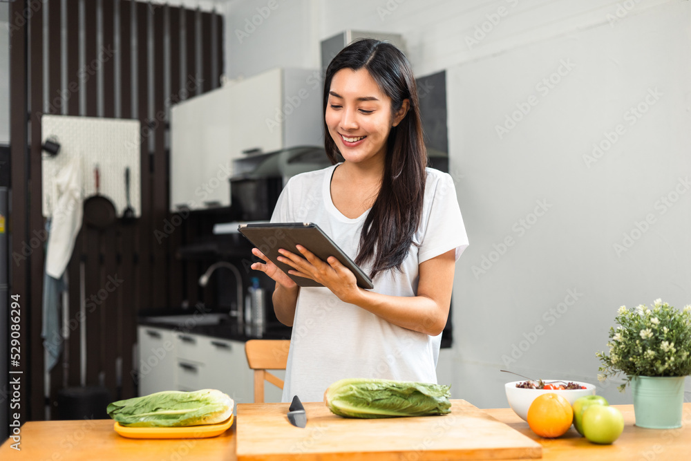 Beautiful woman using tablet to calculate calories and nutrition vegetable salad in weight loss diet. Young female happily cooks a healthy breakfast in kitchen in the morning. Diet food concept.