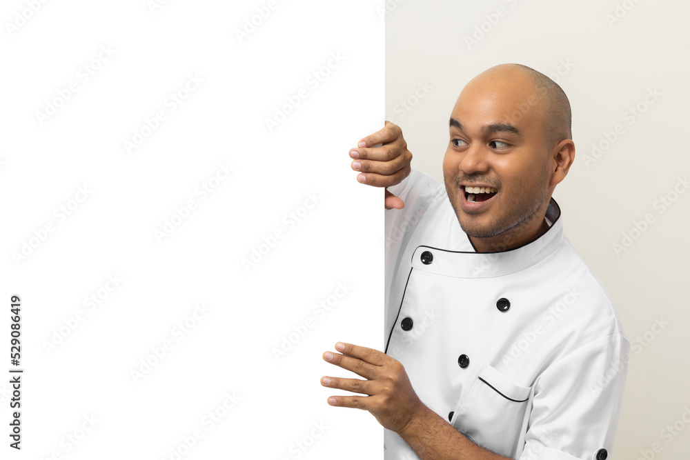 Happy Young handsome asian man chef in uniform looks out from behind an empty whiteboard. Cooking indian man hiding behind big blank billboard for advertising text menu in kitchen and restaurant. Happy Young handsome asian man chef in uniform looks out from behind an empty whiteboard. Cooking indian man hiding behind big blank billboard for advertising text menu in kitchen and restaurant.