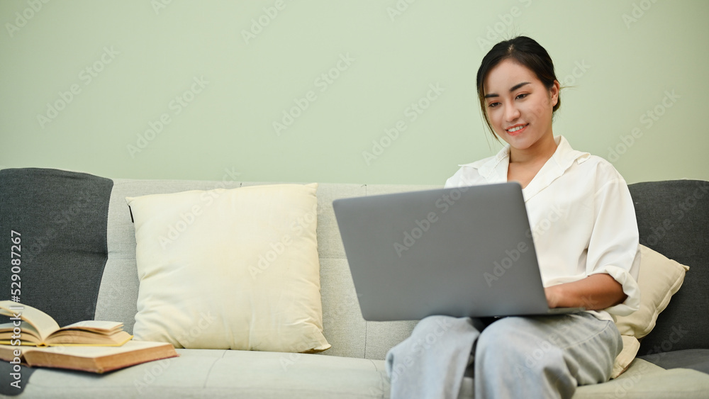 Beautiful Asian female using laptop computer on her comfy sofa in her living room. Beautiful Asian female using laptop computer on her comfy sofa in her living room.
