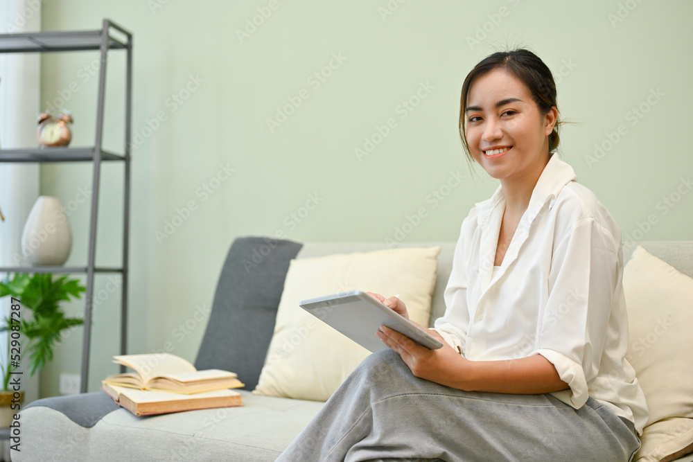 Beautiful and smiling Asian female relaxes in her living room, sits on sofa and uses a tablet. Beautiful and smiling Asian female relaxes in her living room, sits on sofa and uses a tablet.