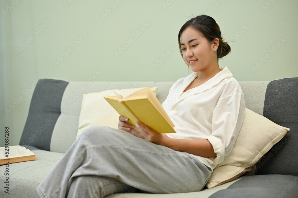 Relaxed and calm Asian woman enjoys reading a book on her cozy sofa in her living room Relaxed and calm Asian woman enjoys reading a book on her cozy sofa in her living room