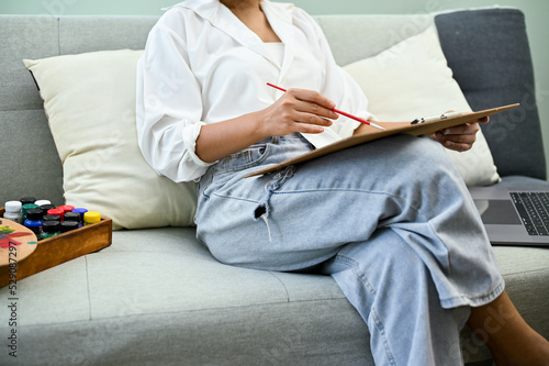 Relaxed Asian female sits on the sofa in her home living room, drawing and painting water color