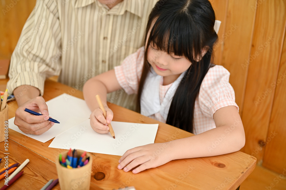 A cute young Asian girl concentrating doing homework on a paper with her dad or private teacher. A cute young Asian girl concentrating doing homework on a paper with her dad or private teacher.