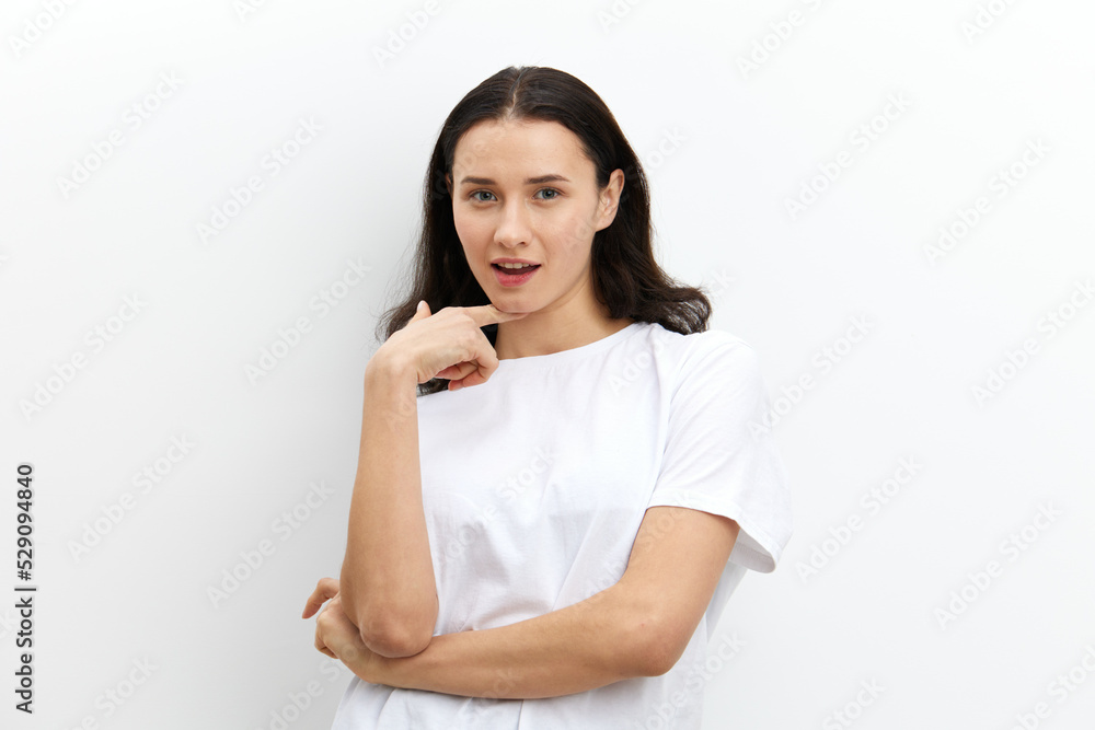 a contented, beautiful, woman with dark, long hair stands on a white background in a light T-shirt and touches her neck with her fingers while looking at the camera a contented, beautiful, woman with dark, long hair stands on a white background in a light T-shirt and touches her neck with her fingers while looking at the camera