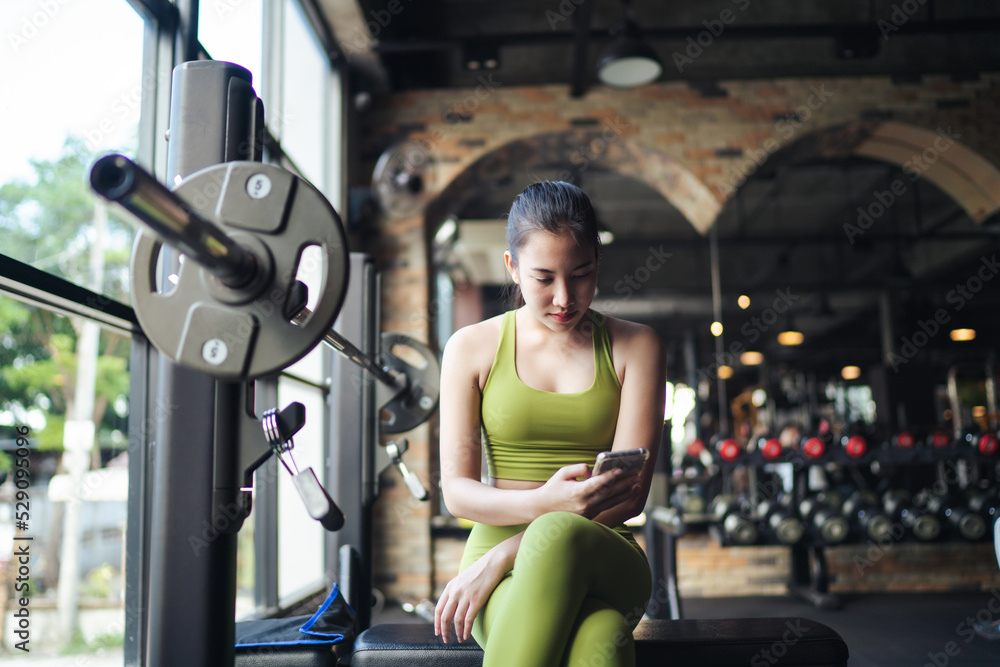 Fototapeta premium Beautiful young Asian woman in sportswear resting or waiting her friend after exercise at fitness gym and using smartphone for communication chatting or social media. Healthy lifestyle.