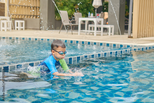 Cute little Asian 6 years old school boy child in swimming goggles and swimming suit learning to swim at outdoor pool in summer day, Happy kid having fun on summer vacation