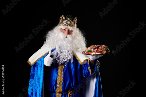 smiling Wise Man holding a "roscón de reyes" in his hand on a black background