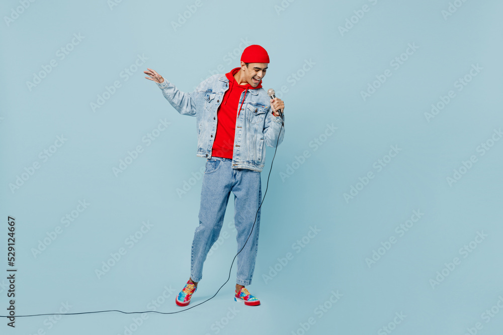 Full body young singer middle eastern man 20s he wear denim jacket red hat sing song in microphone at karaoke club isolated on plain pastel light blue cyan background studio. People lifestyle concept. Full body young singer middle eastern man 20s he wear denim jacket red hat sing song in microphone at karaoke club isolated on plain pastel light blue cyan background studio. People lifestyle concept.