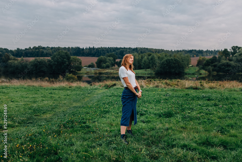 Young woman standing and thinking with a smile on her face. Woman walking at countryside. Woman standing outdoors at cloudy fall day Young woman standing and thinking with a smile on her face. Woman walking at countryside. Woman standing outdoors at cloudy fall day