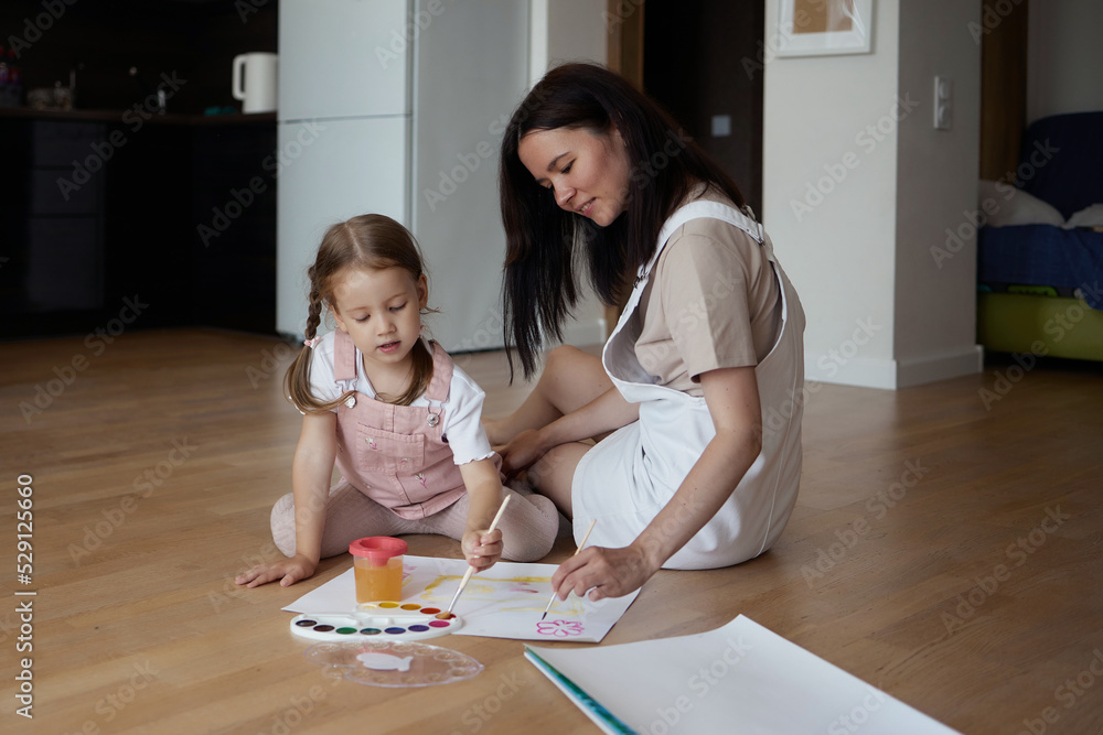 Smiling young mother and little daughter lie on a warm wooden floor in the living room, drawing together, happy mother rejoices at her daughter's drawings. paint on canvas Smiling young mother and little daughter lie on a warm wooden floor in the living room, drawing together, happy mother rejoices at her daughter's drawings. paint on canvas