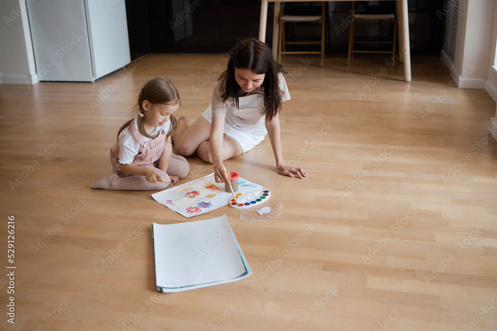 Smiling young mother and little daughter lie on a warm wooden floor in the living room, drawing together, happy mother rejoices at her daughter's drawings. paint on canvas Smiling young mother and little daughter lie on a warm wooden floor in the living room, drawing together, happy mother rejoices at her daughter's drawings. paint on canvas