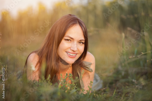 Brown-haired woman lies on grass admiring picturesque countryside landscape. Lady looks around with excited and smiling expression on meadow, sunlight