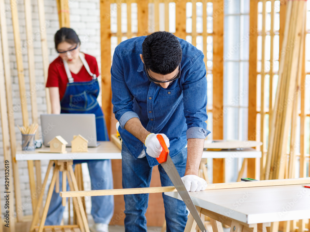 Obraz premium Indian Asian professional male engineer architect foreman labor worker wears safety goggles glasses and gloves using hand saw cutting wood plank on wooden table while female colleague working behind