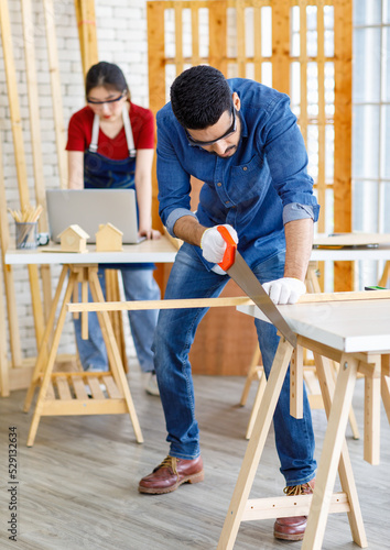 Indian Asian professional male engineer architect foreman labor worker wears safety goggles glasses and gloves using hand saw cutting wood plank on wooden table while female colleague working behind