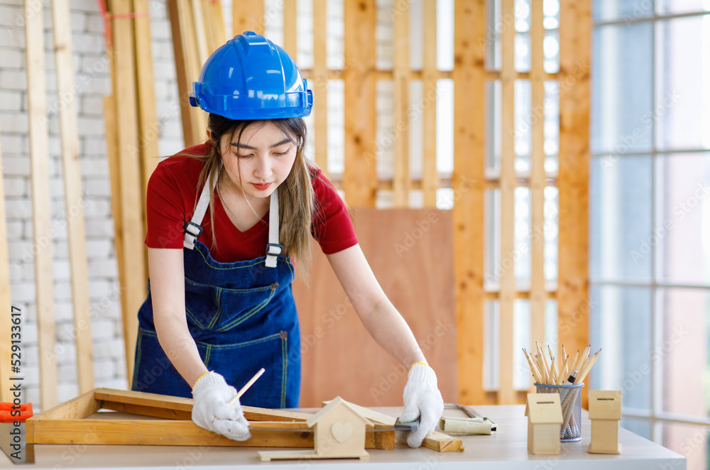 Asian professional female engineer architect foreman labor worker wears safety hard helmet jeans apron and gloves standing using squard angle degree ruler and pencil measuring wood plank on workbench