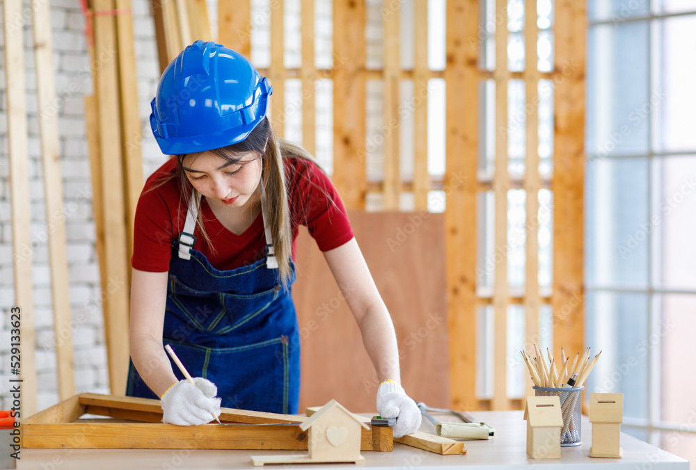 Asian professional female engineer architect foreman labor worker wears safety hard helmet jeans apron and gloves standing using squard angle degree ruler and pencil measuring wood plank on workbench Asian professional female engineer architect foreman labor worker wears safety hard helmet jeans apron and gloves standing using squard angle degree ruler and pencil measuring wood plank on workbench