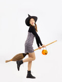 Halloween theme, young asian woman in black dress, boots, witch hat holding broom and carrying orange pumpkin bucket on white background.