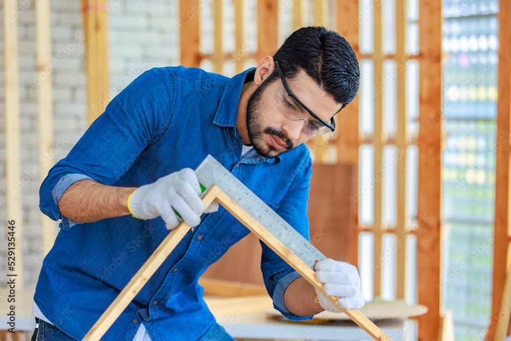 Asian Indian professional male engineer architect foreman labor worker wears safety goggles and gloves using square degree angle steel ruler measuring triangle wood plank in home construction site