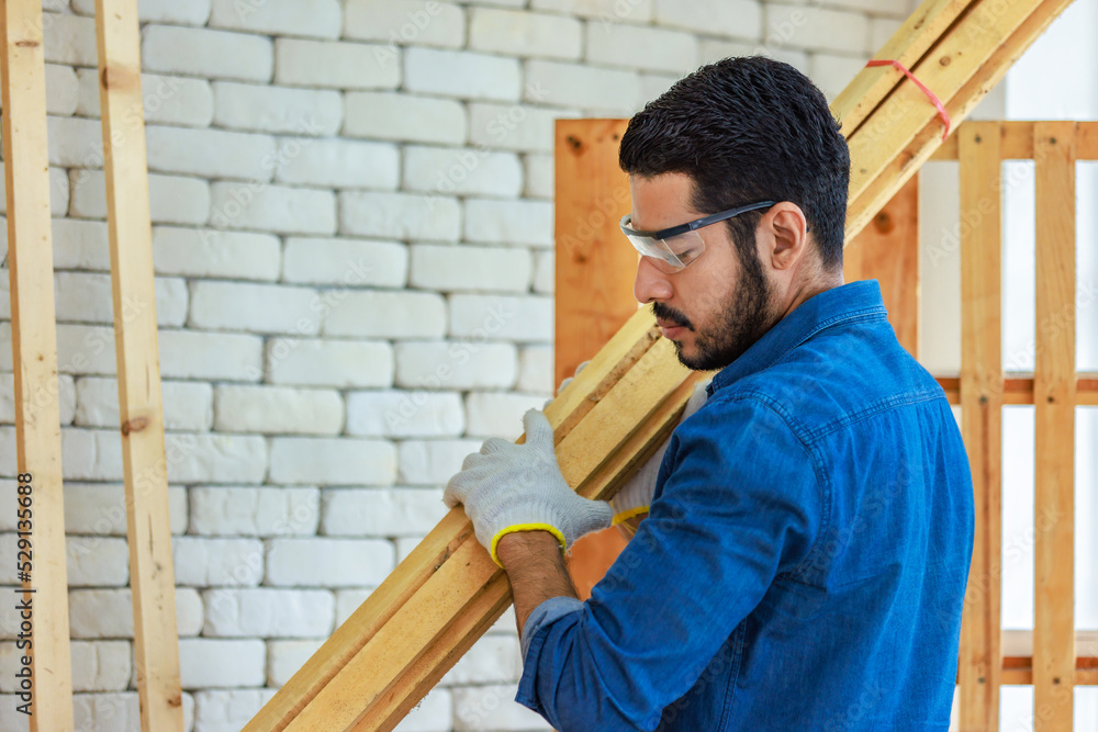 Asian Indian professional bearded male engineer architect foreman labor worker wears safety goggles, hard helmet and gloves standing holding checking wood sticks stock inventory in construction site Asian Indian professional bearded male engineer architect foreman labor worker wears safety goggles, hard helmet and gloves standing holding checking wood sticks stock inventory in construction site